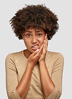 Woman in tan shirt holding her hand to her jaw in pain with white background